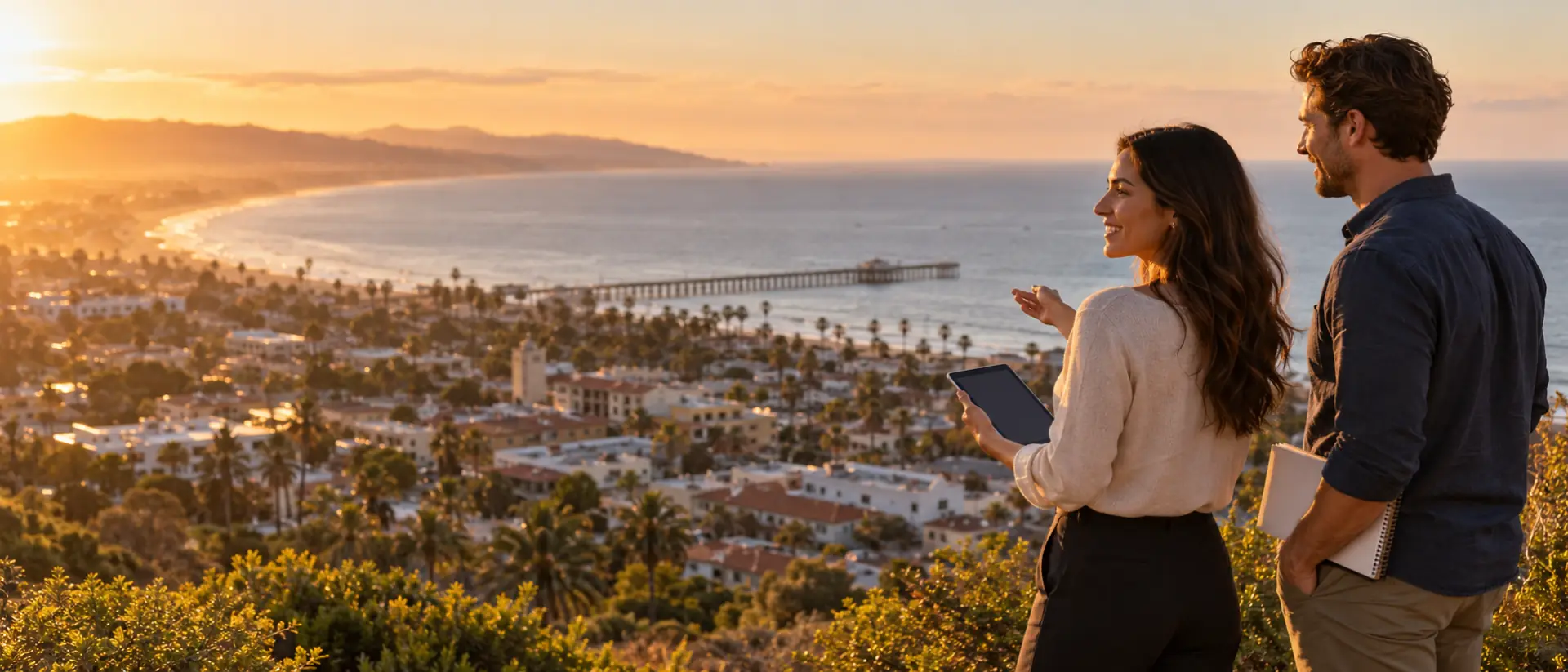 Two marketing professionals with a tablet and notebook at a hillside overlook above downtown Ventura and the pier at golden hour